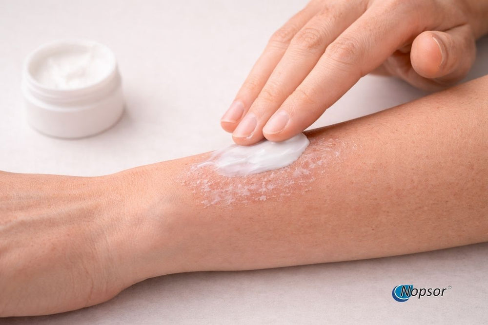 Person applying cream to their leg with a small container of cream beside them on a light background.