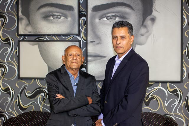 Two men standing in front of a decorative wall with framed portraits.