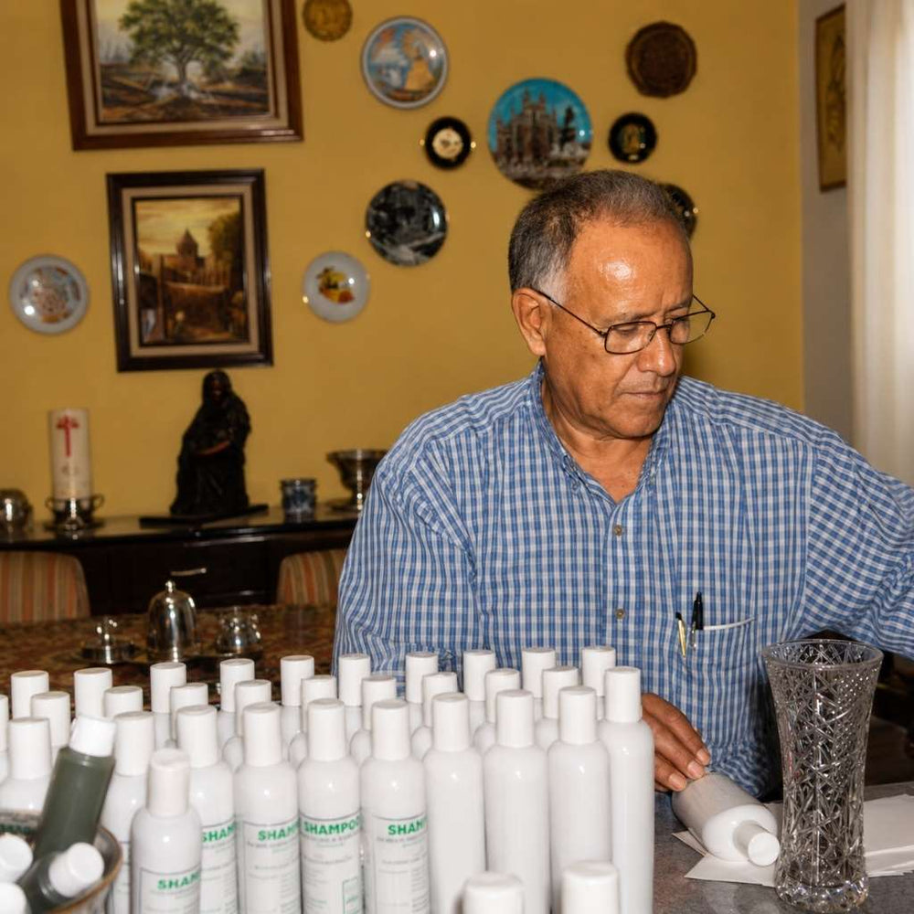 Man in a checkered shirt organizing white bottles with 'Shampoo' labels in a room with decorative plates on the wall.
